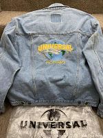 Back view of light blue denim jacket showing large embroidered Universal Pictures logo on back with yellow text over a globe design. Universal plastic bag visible below jacket.