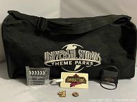 Front side of black Universal Studios Theme Parks tote bag with Universal Studios collectibles arranged in front: keychain, luggage tag, pins, and digital clock.