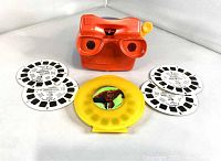 Red 3D Viewmaster viewer with a yellow Incredibles reel holder and six black and white reels arranged around it.