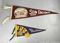 Front view showing both vintage pennants: the long maroon San Antonio Texas pennant with The Alamo illustration and the smaller blue Old Fort Henry Kingston pennant with 1956 and colorful coat of arms.