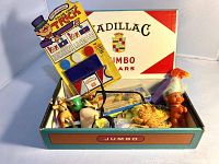 Photo showing assorted toys inside a Jumbo Cigars branded box with Cadillac logos, featuring various plastic small toys and game kit.