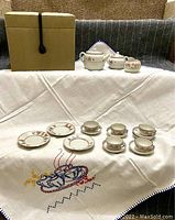 Photo of the tea set pieces arranged on an embroidered tablecloth with the storage box behind