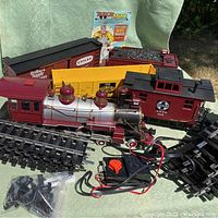 Photo showing train engine, four railroad cars, track sections, wired controller, and instruction booklet on green fabric surface outdoors.