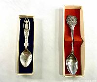 Two souvenir spoons side by side in their original boxes on a white background.