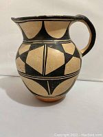 Side view showing the full body of the clay pitcher with black geometric decoration and beige background