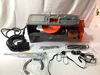 Overview of the Champion Wheelybird electronic clay target thrower with included parts arranged in front of it: foot pedal, metal rods, springs, clamps, and wiring harness.