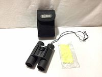 Photo shows Bushnell-branded carrying case, 12x25 binoculars with neck strap, and a yellow cleaning cloth in plastic wrapping.