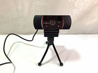 Front view of Thronmax StreamGo webcam mounted on small tripod with USB cable connected, showing the camera lens and noise-reducing microphone grills on both sides.