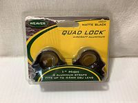 Front of product package showing Weaver Quad Lock Mount in matte black, specifying 1 inch high and 4 aluminum straps fit for up to 44mm objective lenses.