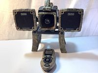 Front view of Primos Boss Dogg Predator Caller with two rectangular speakers and central horn speaker on camo legs, and front-facing remote control with TFT screen and control buttons.