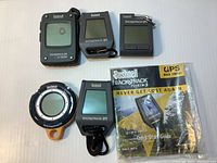 Five Bushnell BackTrack GPS devices arranged on white surface with Quick Start Guide booklet. Top three devices have active display screens, bottom two devices do not power on. Devices vary in shape including rectangular and circular design, color dark grey with some orange on circular device.