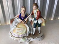 Front view of porcelain figurine showing woman seated and man standing playing lute, set on ornate base.