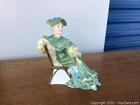Side angle showing seated woman figurine in green dress and hat with parasol on chair