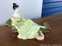 Side angled view of Royal Doulton figurine showing woman's green dress, chaise lounge, and fruit basket.