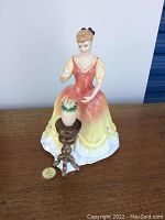 Front view of Royal Doulton porcelain figurine "Sarah" seated beside a wooden style side table holding a flower vase with colorful flowers.