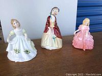 Front view of all three Royal Doulton figurines on a wooden surface: Paisley Shawl in the center, January left, Rose right.