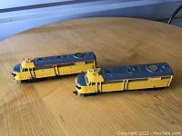 Two yellow and gray Union Pacific model train engines on a wooden surface, showing side view and front details.