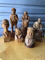 Seven wooden carvings on a table: four full figures and three busts, showing men in various poses and attire, each approximately 18 inches tall.