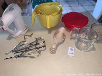 Full lot of baking and kitchen items arranged on table: white Oster hand mixer, yellow Rubbermaid mixing bowls nested, glass measuring cup with red lid, Ekco measuring spoons, and mixer attachments.