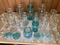 Front view of 17 glass jars with 12 glass lids arranged in front on a white cloth. Mix of clear and blue-tinted glass jars, some with wire bale handles.