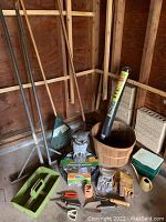 Wide view of the garden tools arranged in shed corner showing long-handled tools, owl statue, basket, edging rolls, gloves and small pots.