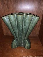 Fan-shaped, footed vase with green and blue speckled glaze shown from frontal angle against wooden background.