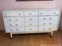 Front view of the white painted dresser showing nine drawers with floral enamel knobs and tapered legs