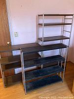 Three metal shelving units: two silver frame shelves with dark wood-look adjustable shelves, and one black metal shelf with silver cylinder sides.