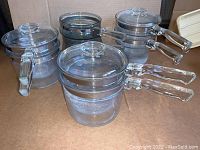 Group photo showing six clear glass Pyrex double boilers with handles and lids, arranged on a brown surface.