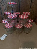 Group of 13 vintage Smuckers jelly jars stacked and arranged on a cardboard surface, showing clear glass jars with embossed branding and red and white checkered lids.