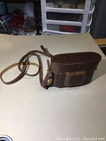 Brown leather carrying case with strap, showing minor wear and embossed logo mark on front.