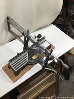 Front angle showing the full miter saw mounted on a wooden base with visible adjustable angle gauge and clamp mechanism