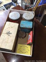Multiple different metal tobacco tins and boxes of various shapes including John Player tobacco box with empty cans inside, red rectangular tin, blue square tin, cylindrical tin, and a yellow 555 BOAC tobacco tin.