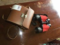 Binoculars and brown leather carrying case with strap and clasp placed on wooden surface.