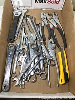 Box containing assorted wrenches, two pliers, ratchet wrench, and yellow box cutter showing wear and usage marks.