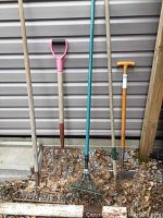Wide view of five yard tools leaning against a siding wall, showing entire tools with handles and heads.