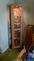 Front view of tall narrow wooden cabinet with glass door showing shelves and interior light illuminating collection inside.