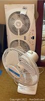 Both fans shown, large SMC window fan behind smaller white Holmes tabletop fan on carpeted floor, visible power cords