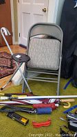 Two gray metal folding chairs with padded striped fabric seats, a folding sport cane with a small round black seat, several umbrellas, flashlights and tools placed on carpet floor near a closed white door.