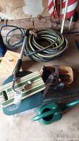 Coiled garden hose, gutter cleaner attachment, folding garden seat with cushions, wooden toolbox containing hand tools, and woven basket arranged on the floor.