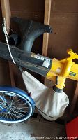 Side view of yellow Yard-Man blower/vacuum with attached beige collection bag and black nozzle attachments hanging on wall.