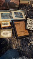 Photo showing multiple framed items, including metallic art, vintage prints, public sale poster, and framed collection of keys placed on a carpeted floor.