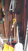 Photo of various snow shovels, garden claw, and hand tools leaning against a wooden wall.