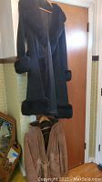 Black suede long coat hanging on door, showing shearling trim on cuffs and collar.