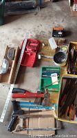 Photo showing assorted hand tools laid out on concrete floor including saws, mitre box, hammer, socket set with bits, nails, and yard stick.