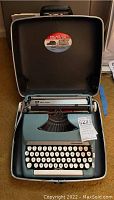 Blue Smith Corona Sterling typewriter inside black hard case with lid open, shows keyboard and writing roller.