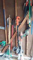 Photo showing yard tools leaning against garage wall including Black and Decker orange electric trimmer and edger, rakes, shovel, and hand tools