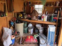 Wooden garden shed shelf with bird feeder, plant pots, red mulch bag, and various gardening items.