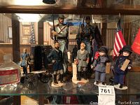 Wide view of Civil War figurines, dolls, flags, and cannon model arranged on shelf.