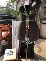 Full view of adjustable multi-piece dress form torso on metal stand, visible outdoors near other furniture items, tagged with item number label.